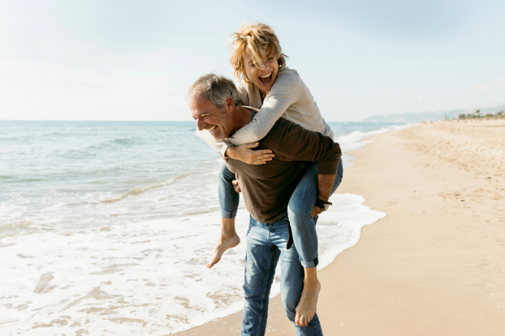 Couple enjoying a playful moment on the beach, with the man giving a piggyback ride to the woman, symbolizing joy and connection in a wellness-focused lifestyle.