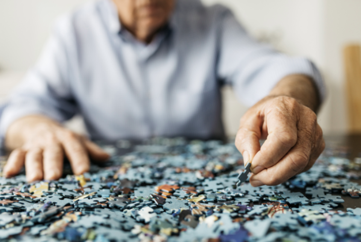 Elderly person assembling a jigsaw puzzle, focusing on a blue piece among a colorful assortment, illustrating cognitive engagement and leisure activity.